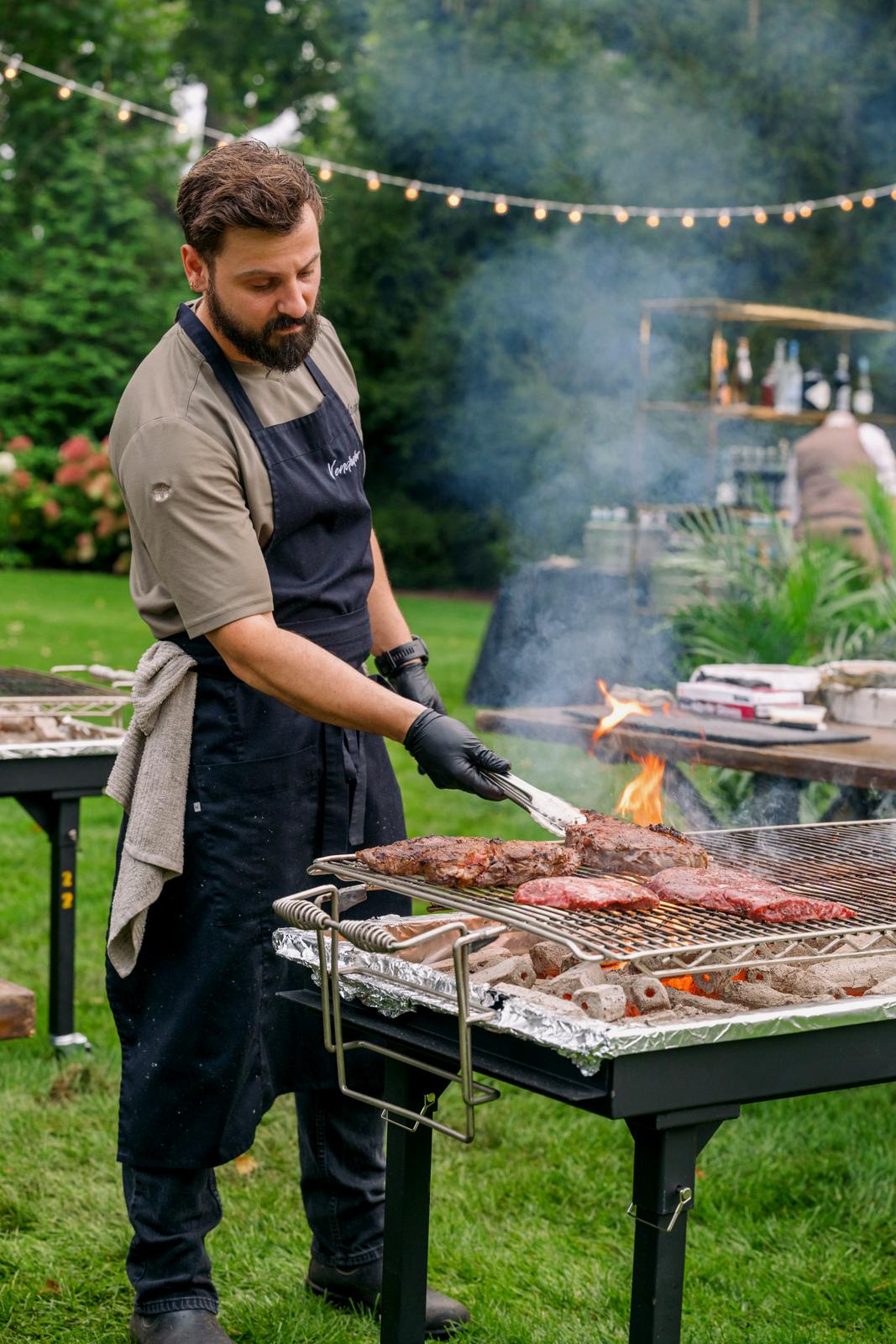 A Vendador chef grilling steaks over live fire in the garden