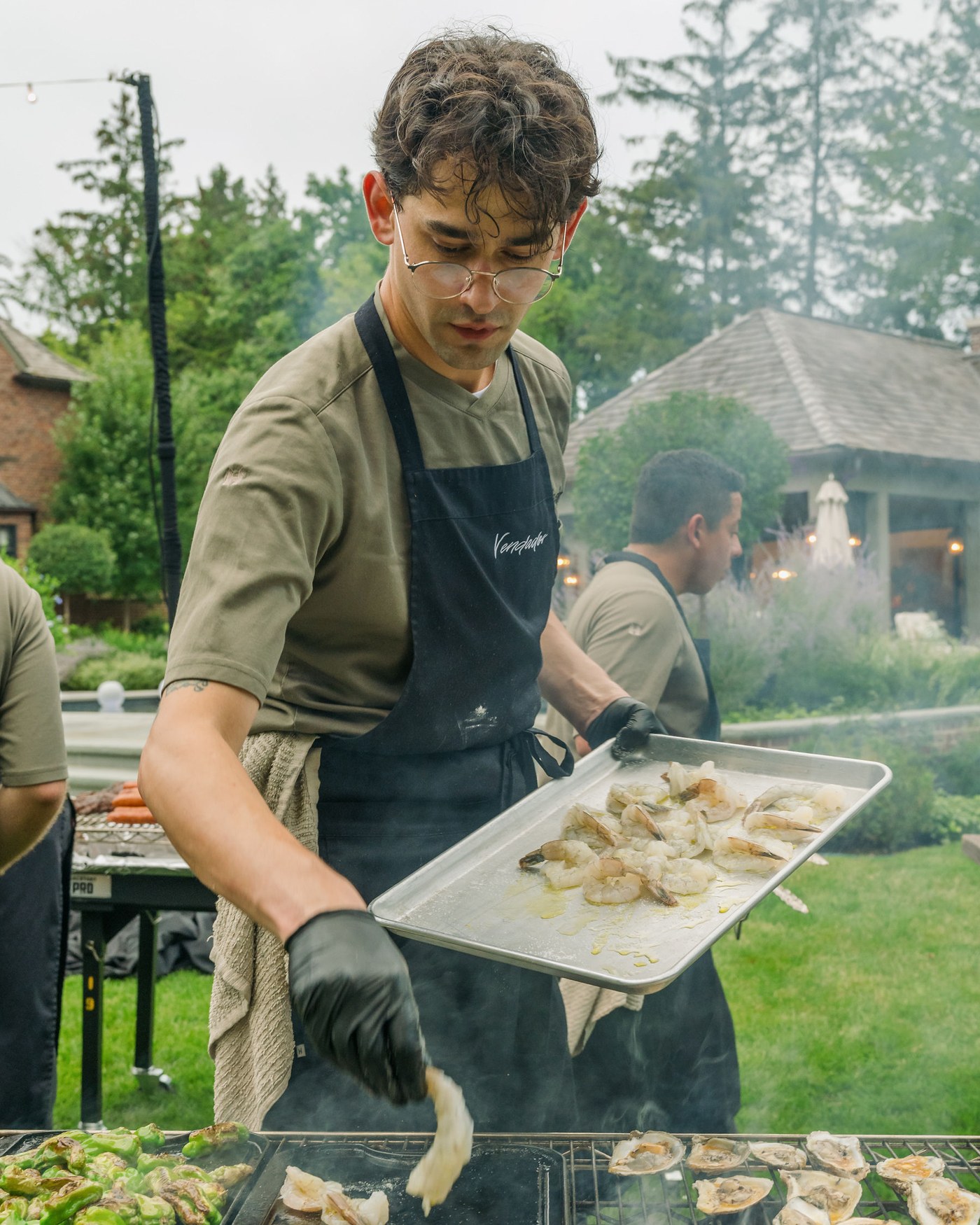 A Vendador chef laying shrimp on the grill beside shishitos and oysters